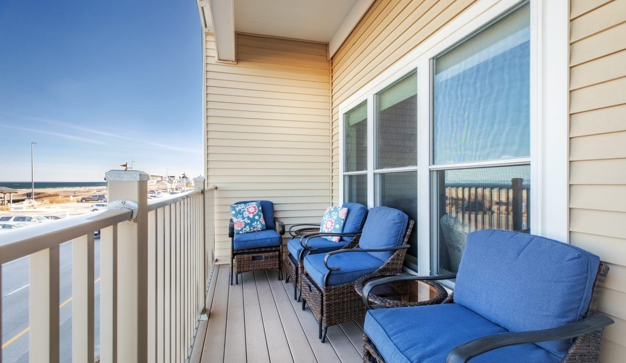 A balcony with wicker chairs featuring blue cushions and floral pillows, a small table, beige siding, and a view of the street and water in the background.