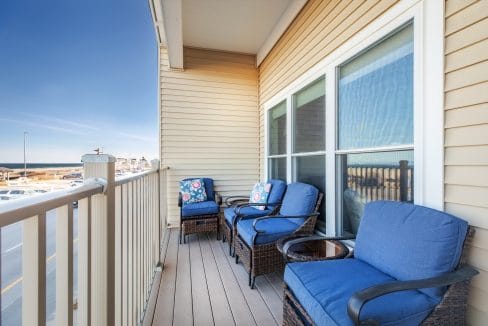 A balcony with wicker chairs featuring blue cushions and floral pillows, a small table, beige siding, and a view of the street and water in the background.