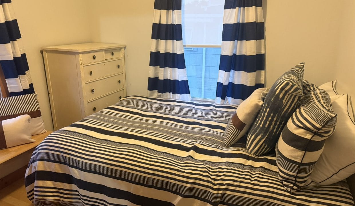 A bedroom with a bed covered in blue and white striped bedding, matching curtains, several pillows, a white dresser, and a small wooden bench.