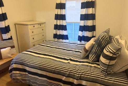 A bedroom with a bed covered in blue and white striped bedding, matching curtains, several pillows, a white dresser, and a small wooden bench.
