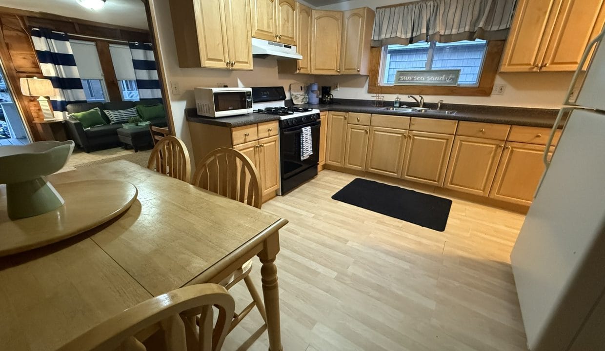 A kitchen with light wood cabinets, a dining table with chairs, a white refrigerator, stove, microwave, and a window with curtains; a living room is visible in the background.