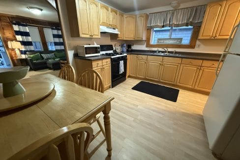A kitchen with light wood cabinets, a dining table with chairs, a white refrigerator, stove, microwave, and a window with curtains; a living room is visible in the background.