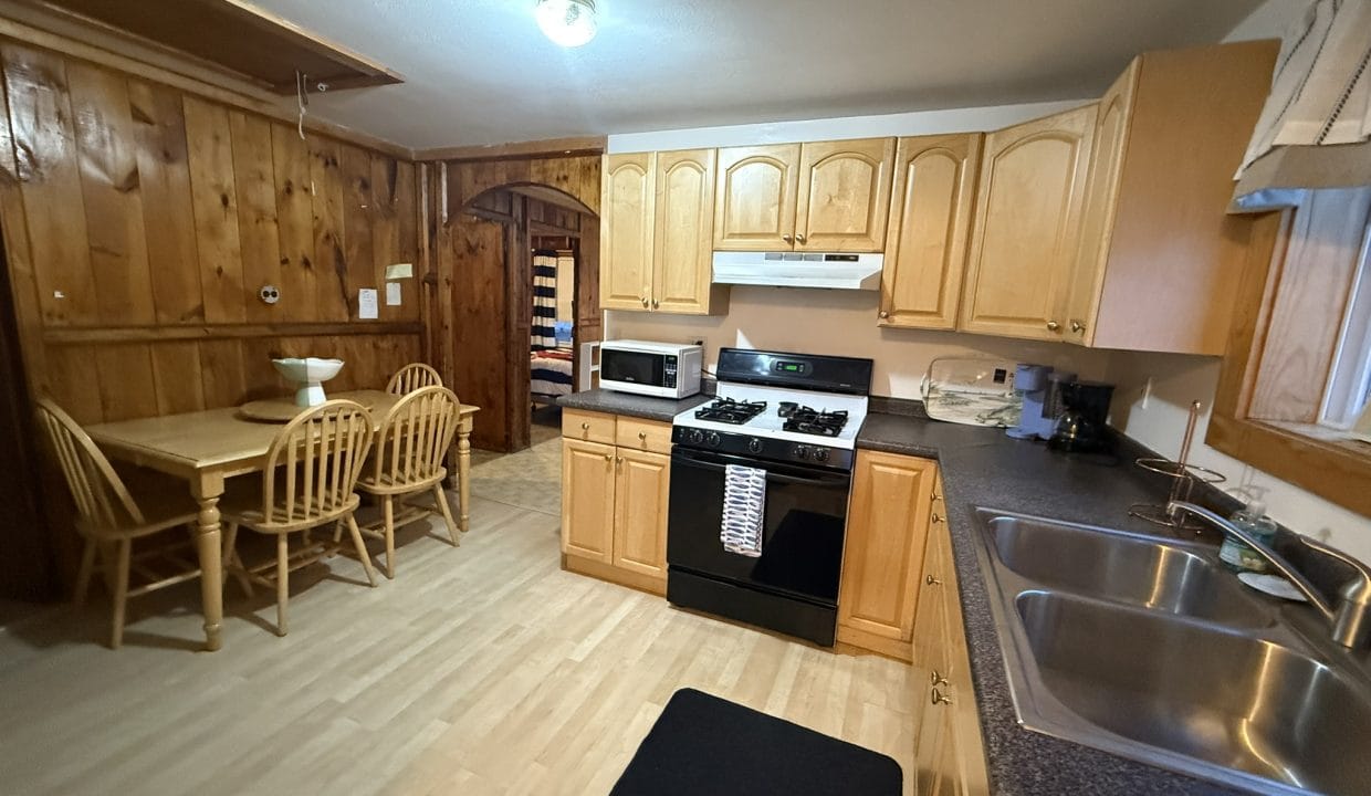 Kitchen with wood-paneled walls, light wood cabinets, a black stove, microwave, double sink, and a small dining table with four chairs.
