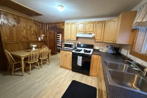 Kitchen with wood-paneled walls, light wood cabinets, a black stove, microwave, double sink, and a small dining table with four chairs.