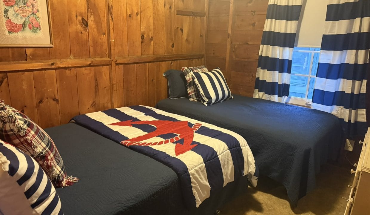 A small wooden-paneled bedroom with two twin beds, navy and white striped bedding, plaid accent pillows, and a window with matching striped curtains.