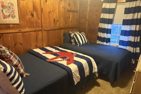 A small wooden-paneled bedroom with two twin beds, navy and white striped bedding, plaid accent pillows, and a window with matching striped curtains.