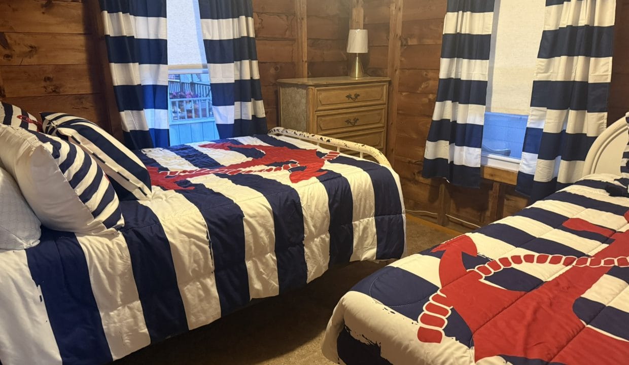 Two beds with navy blue and white striped bedding and red anchor designs in a wood-paneled room, with a dresser and two windows with matching curtains.