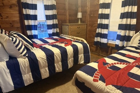 Two beds with navy blue and white striped bedding and red anchor designs in a wood-paneled room, with a dresser and two windows with matching curtains.