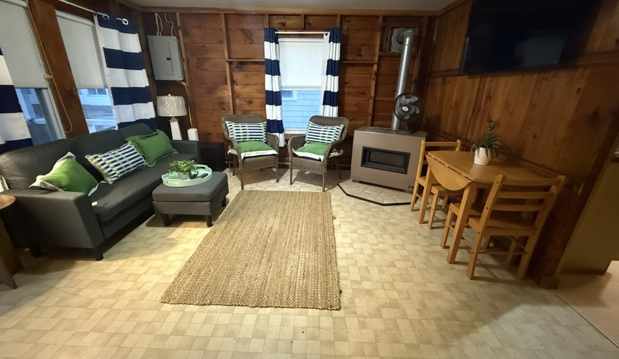 A cozy living room with wood-paneled walls, a black sofa, two chairs, a jute rug, a small stove, dining table with chairs, and striped curtains on the windows.
