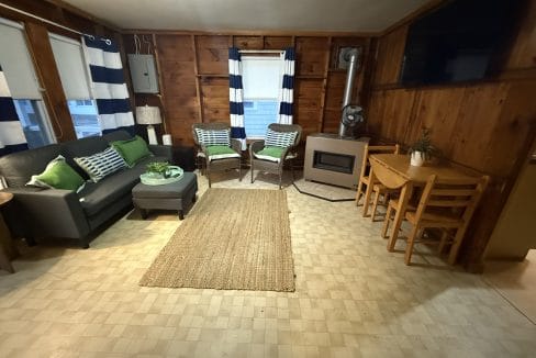 A cozy living room with wood-paneled walls, a black sofa, two chairs, a jute rug, a small stove, dining table with chairs, and striped curtains on the windows.