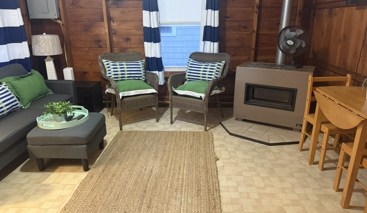 A cozy room with wood-paneled walls, striped curtains, wicker chairs, a gray sofa, a wood stove, a fan, and a small wooden dining table with two chairs.
