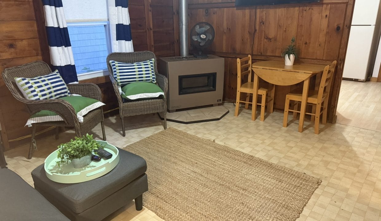 A cozy living area with wicker chairs, a small dining table, a fireplace, a TV, a refrigerator, and a woven rug on a tiled floor.