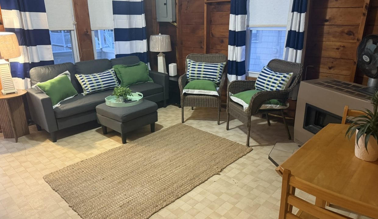 A cozy living room with a black loveseat, two wicker chairs, a rug, a small table with chairs, and blue-striped curtains on wood-paneled walls.