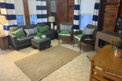 A cozy living room with a black loveseat, two wicker chairs, a rug, a small table with chairs, and blue-striped curtains on wood-paneled walls.