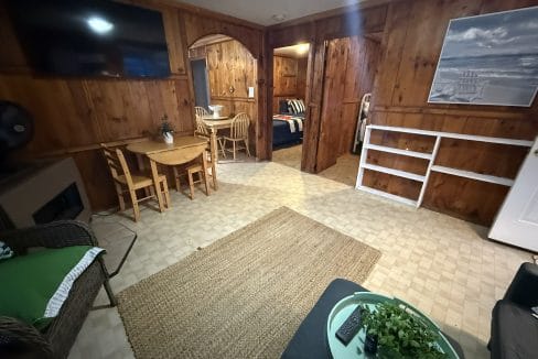 A cozy, wood-paneled living area with a rug, table and chairs, TV, and adjoining bedroom in the background.
