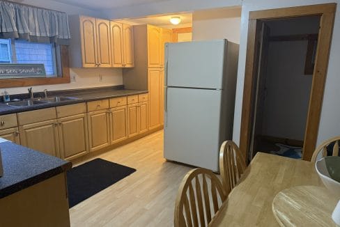 A kitchen with light wood cabinets, white refrigerator, dark countertops, a sink under a window, and a wooden dining table with four chairs.