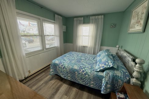 A small bedroom with teal walls, wood flooring, a double bed with blue patterned bedding, two windows with white curtains, and a wooden nightstand.