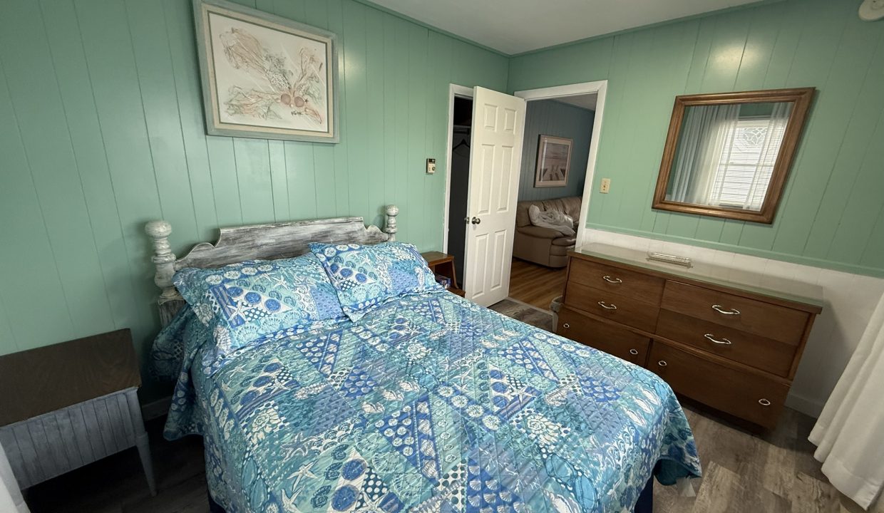 Bedroom with teal walls, a bed with blue-patterned bedding, a dresser with a mirror, a framed picture above the bed, and a partially open door revealing another room.