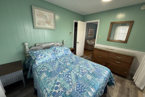 Bedroom with teal walls, a bed with blue-patterned bedding, a dresser with a mirror, a framed picture above the bed, and a partially open door revealing another room.