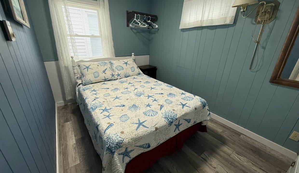 A small bedroom with blue paneled walls, a bed with a starfish-patterned quilt, a window with sheer curtains, a wall lamp, and an open shelf with hangers.