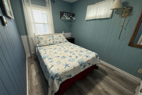 A small bedroom with blue paneled walls, a bed with a starfish-patterned quilt, a window with sheer curtains, a wall lamp, and an open shelf with hangers.