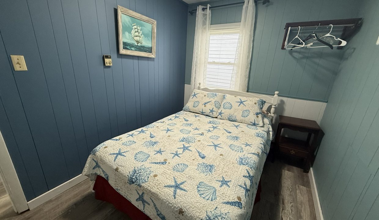 Small bedroom with blue paneled walls, a bed with a seashell-print quilt, a window with sheer curtains, a wall-mounted ship painting, and an open rack with hangers.