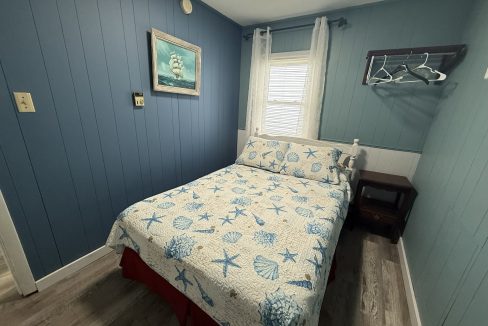 Small bedroom with blue paneled walls, a bed with a seashell-print quilt, a window with sheer curtains, a wall-mounted ship painting, and an open rack with hangers.