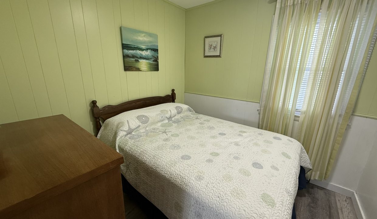 A small bedroom with light green walls, a bed with a patterned quilt, a wooden dresser, two wall hangings, and a window with cream-colored curtains.