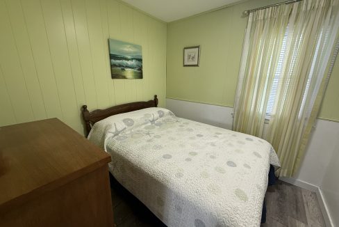A small bedroom with light green walls, a bed with a patterned quilt, a wooden dresser, two wall hangings, and a window with cream-colored curtains.