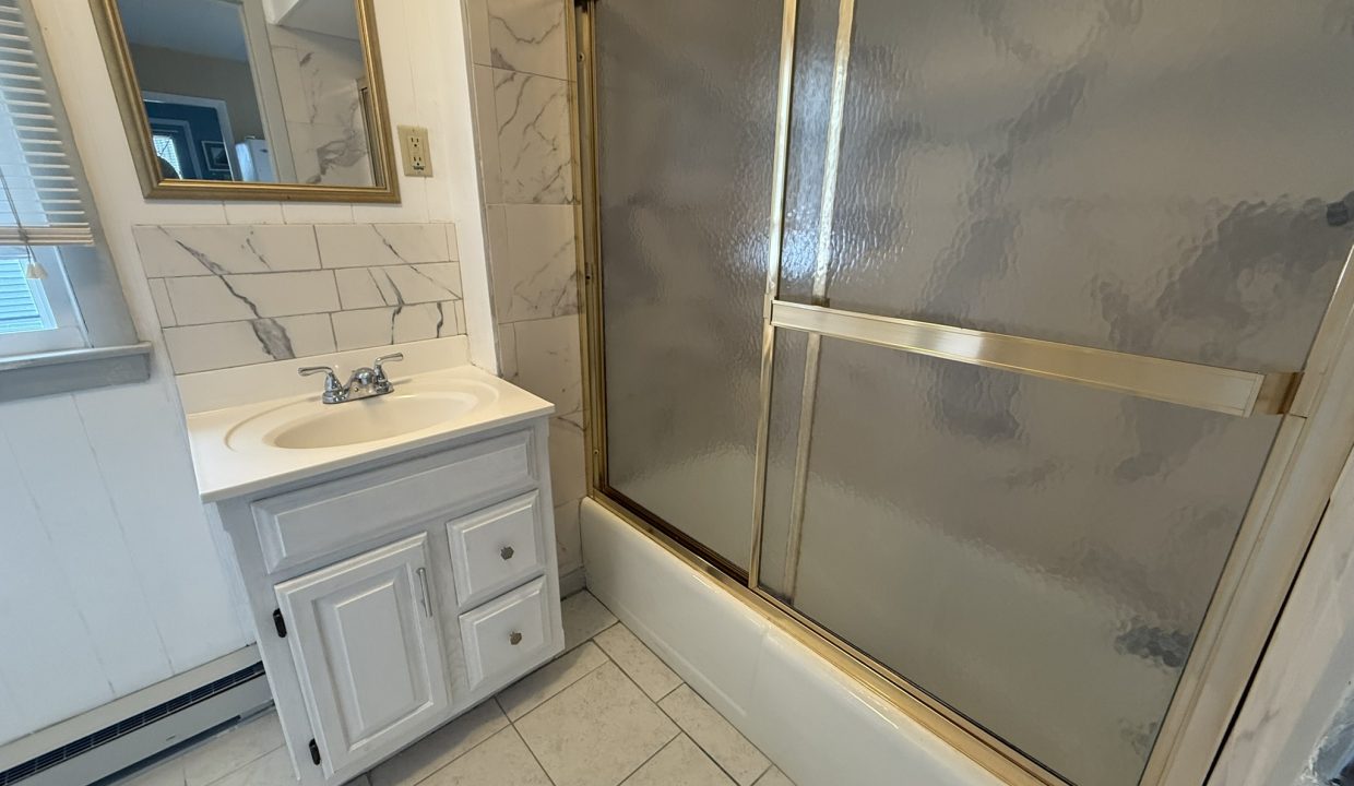 Small bathroom with a white vanity, wall mirror, tiled floor, marble-look wall tiles, and a bathtub with a sliding glass shower door framed in gold.