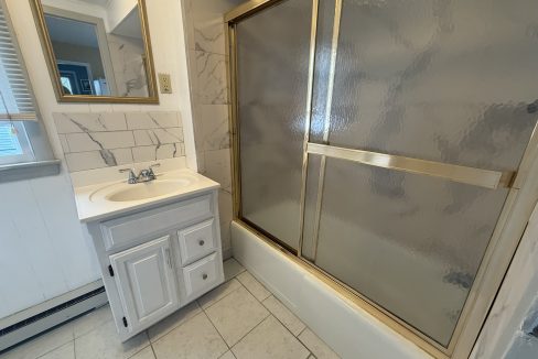 Small bathroom with a white vanity, wall mirror, tiled floor, marble-look wall tiles, and a bathtub with a sliding glass shower door framed in gold.