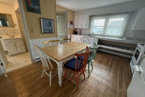 A small kitchen with a wooden dining table and five mismatched chairs, white cabinets, large windows, and an adjacent bathroom visible through an open door.