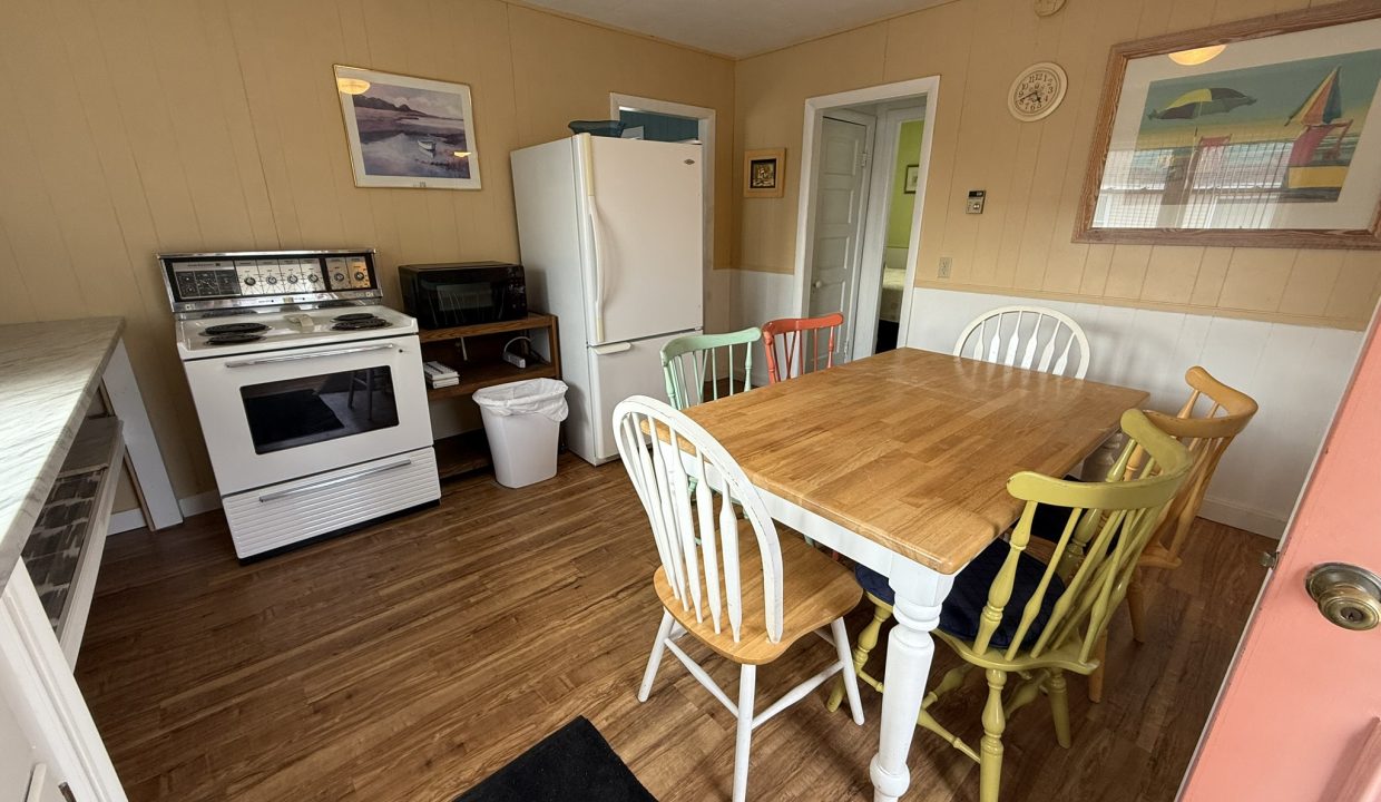 A small kitchen with a white stove, refrigerator, microwave, and a wooden dining table surrounded by mismatched colorful chairs. Paintings hang on the beige walls.