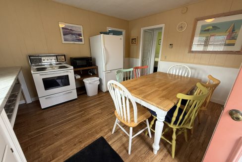 A small kitchen with a white stove, refrigerator, microwave, and a wooden dining table surrounded by mismatched colorful chairs. Paintings hang on the beige walls.
