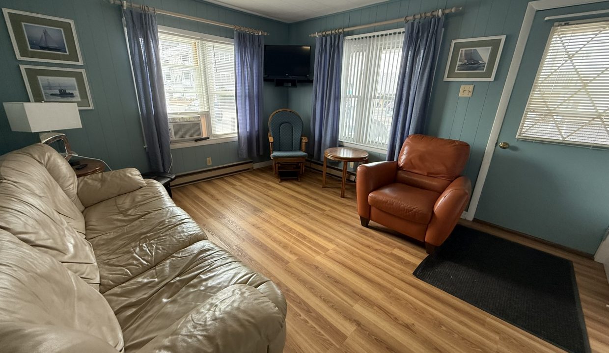 A small living room with a beige sofa, a brown armchair, wood flooring, blue walls, two windows with blue curtains, and a TV mounted in the corner.