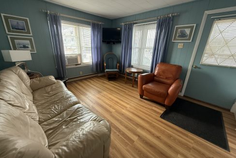 A small living room with a beige sofa, a brown armchair, wood flooring, blue walls, two windows with blue curtains, and a TV mounted in the corner.