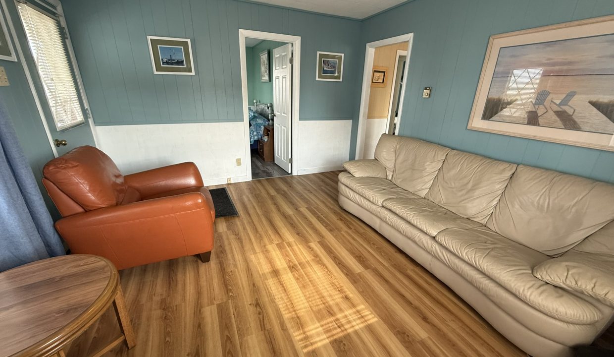 A living room with teal walls, wood flooring, a tan sofa, an orange armchair, a round wooden table, and wall art featuring a beach scene.