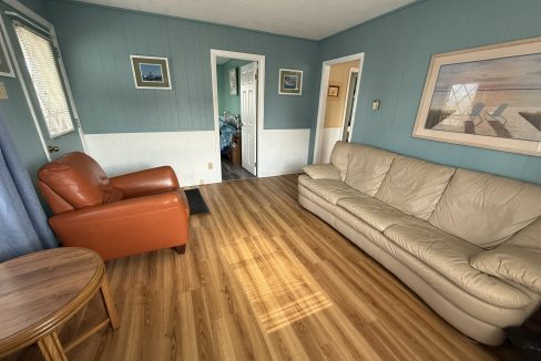 A living room with teal walls, wood flooring, a tan sofa, an orange armchair, a round wooden table, and wall art featuring a beach scene.