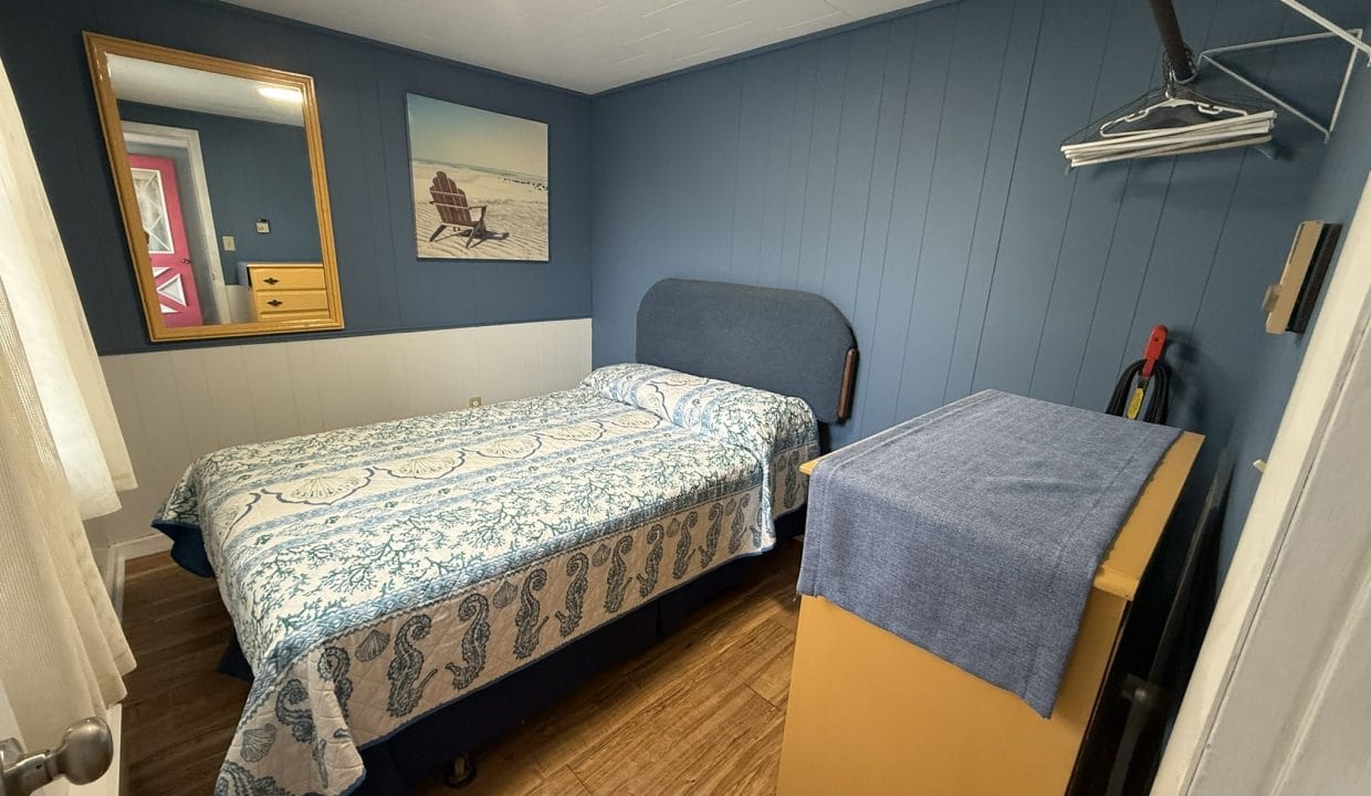 Small bedroom with blue walls, a single bed with patterned bedding, a dresser with a blue cover, wall art, a mirror, and an open shelf with hangers.