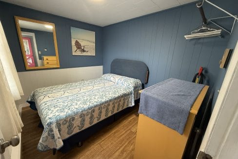 Small bedroom with blue walls, a single bed with patterned bedding, a dresser with a blue cover, wall art, a mirror, and an open shelf with hangers.