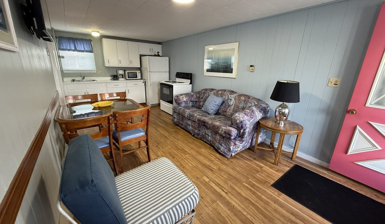 Small living space with a kitchenette, floral sofa, striped chair, wooden dining table, and a pink door on wood flooring.