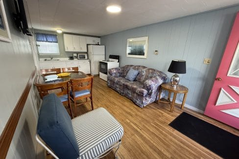 Small living space with a kitchenette, floral sofa, striped chair, wooden dining table, and a pink door on wood flooring.
