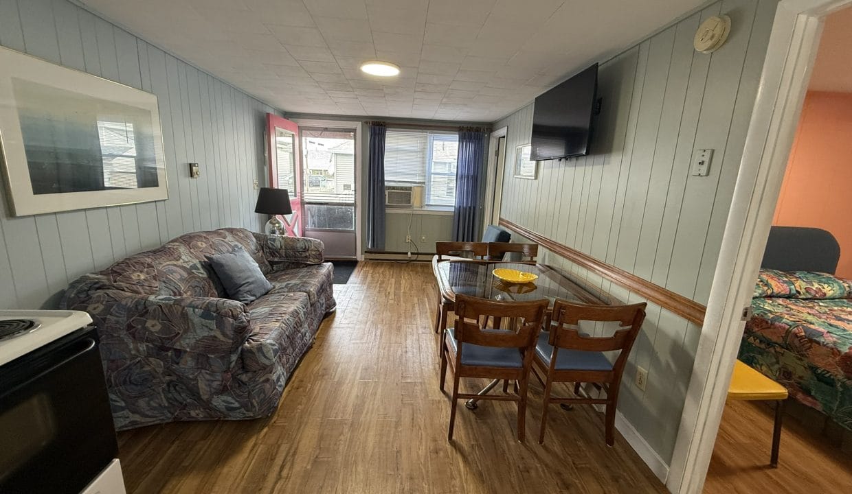 A small living area with a patterned sofa, a wall-mounted TV, a glass dining table with chairs, and a view into a bedroom with an orange wall and bed.