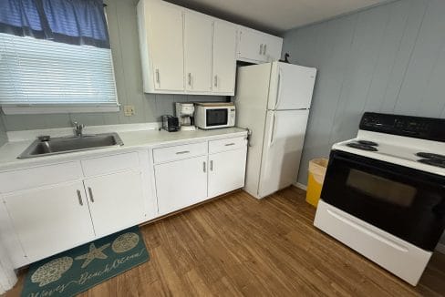 Small kitchen with white cabinets, refrigerator, stove, microwave, coffee maker, and sink. Wooden floor and a window with blue curtained valance above the sink.