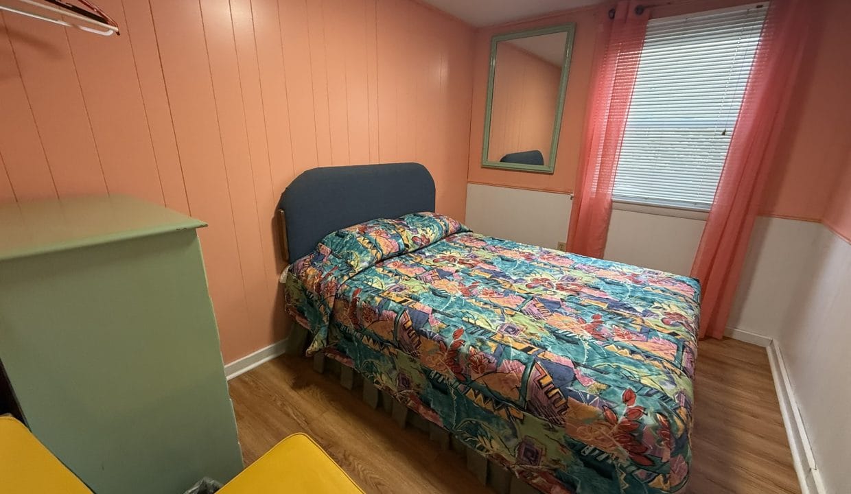 Small bedroom with peach-colored walls, a bed covered in a colorful floral bedspread, a green dresser, two yellow chairs, a wall mirror, and a window with pink curtains.