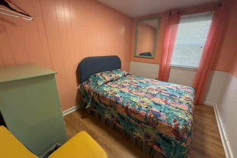 Small bedroom with peach-colored walls, a bed covered in a colorful floral bedspread, a green dresser, two yellow chairs, a wall mirror, and a window with pink curtains.