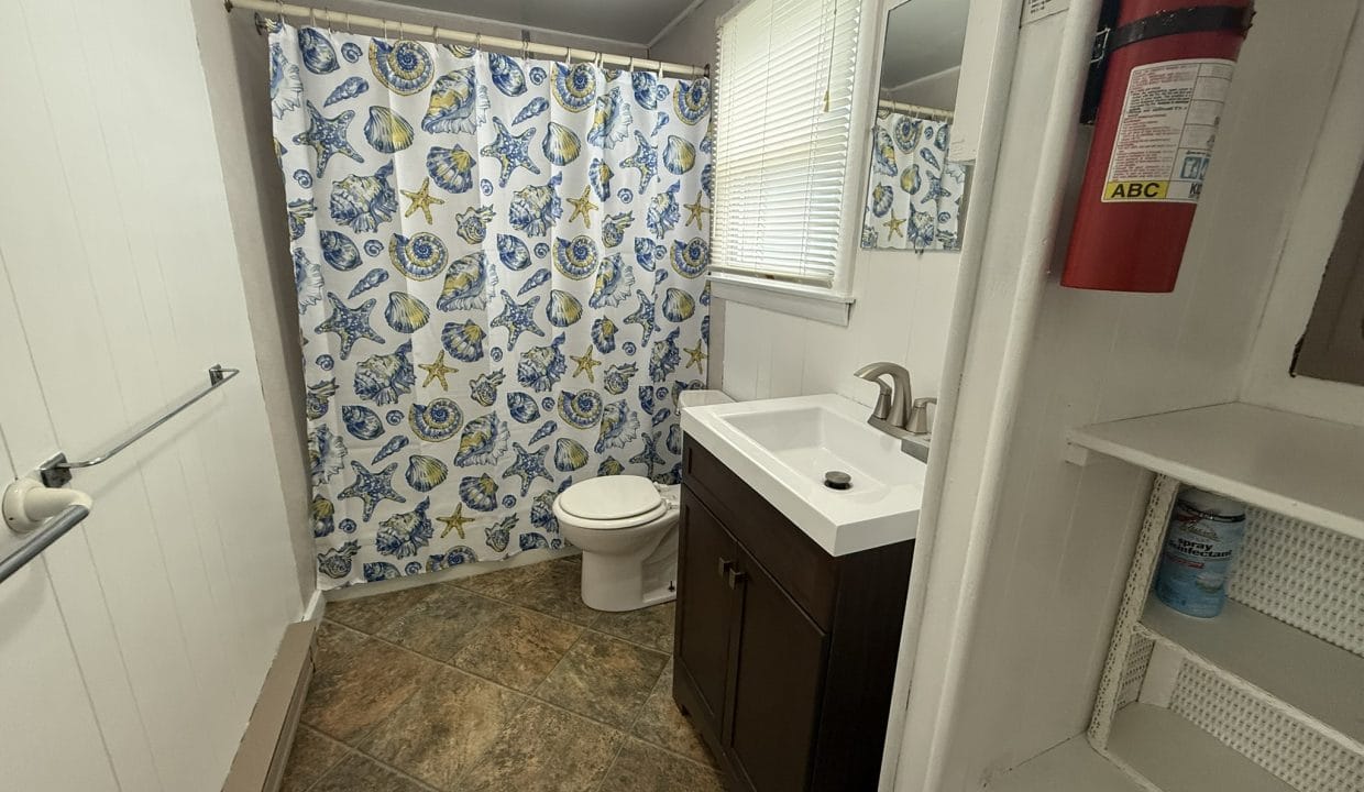Small bathroom with a toilet, sink, floral shower curtain, tiled floor, window with blinds, fire extinguisher on the wall, and open shelves with cleaning products.
