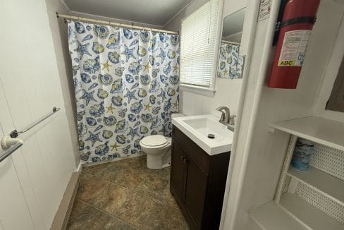 Small bathroom with a toilet, sink, floral shower curtain, tiled floor, window with blinds, fire extinguisher on the wall, and open shelves with cleaning products.