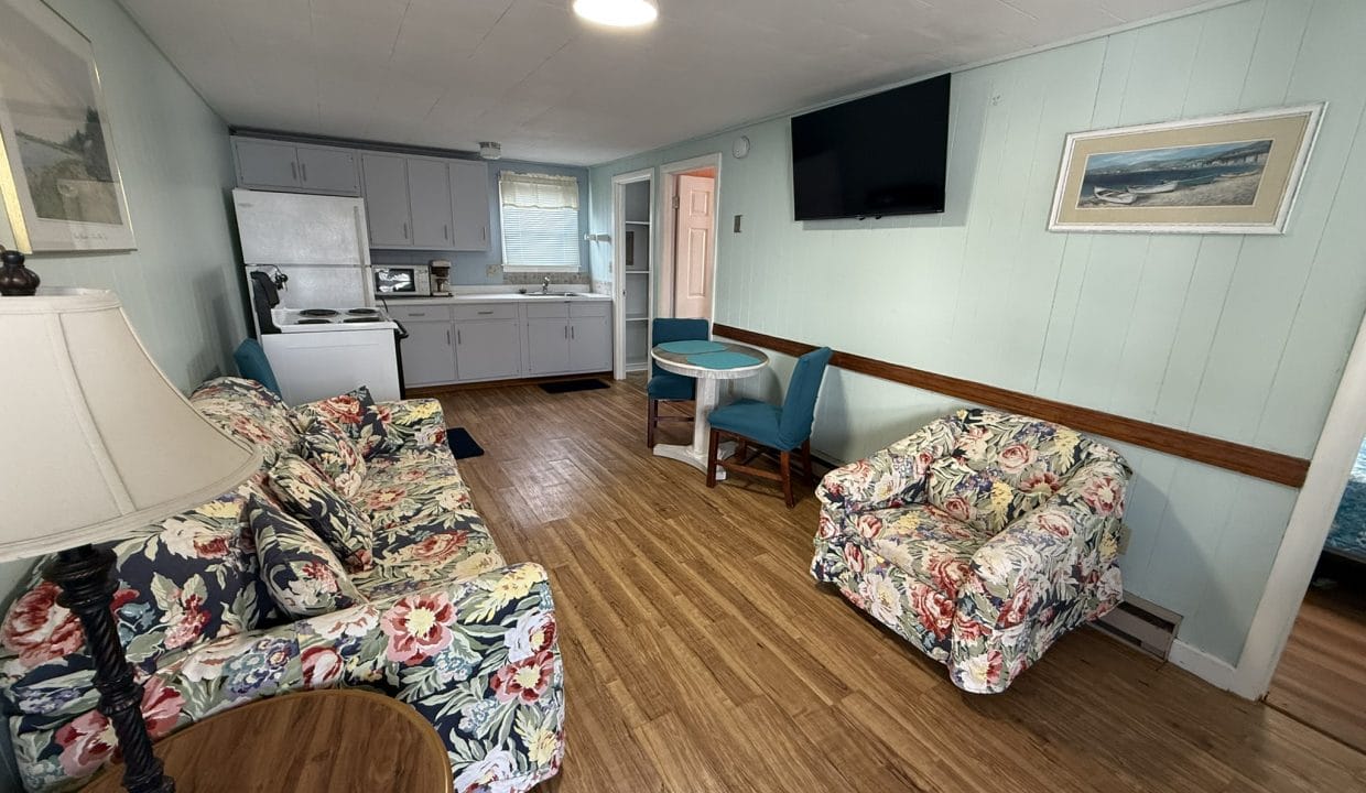 Small living area with floral-patterned couch and armchair, a round table, TV on the wall, and an open view of the kitchen with white cabinets and appliances.