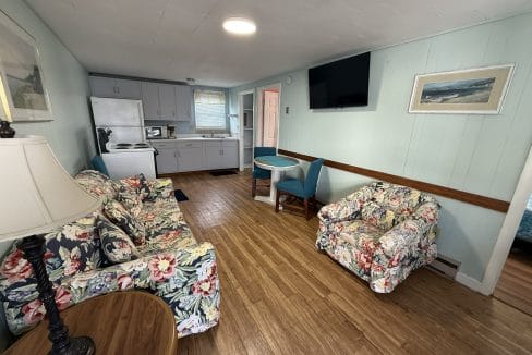 Small living area with floral-patterned couch and armchair, a round table, TV on the wall, and an open view of the kitchen with white cabinets and appliances.